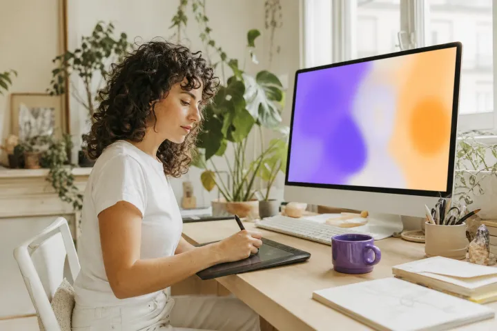 Une jeune femme dessine sur une tablette graphique devant un grand écran aux formes colorées.