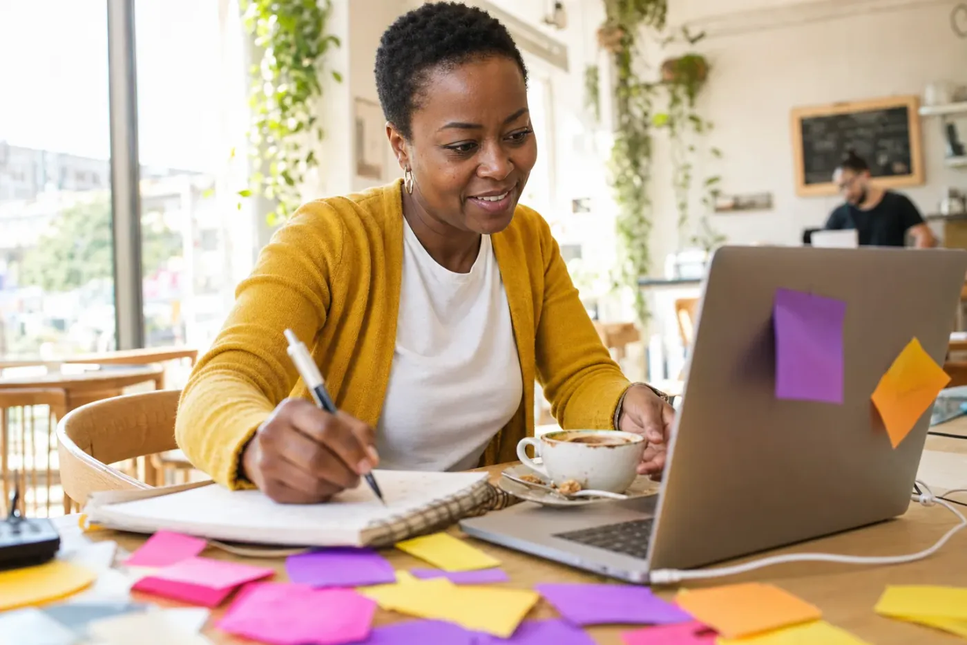 Dans un café inondé de soleil, une femme sourit en notant sa demande dans un carnet avant de l’écrire sur son portable, des post-it colorés autour d’elle.
